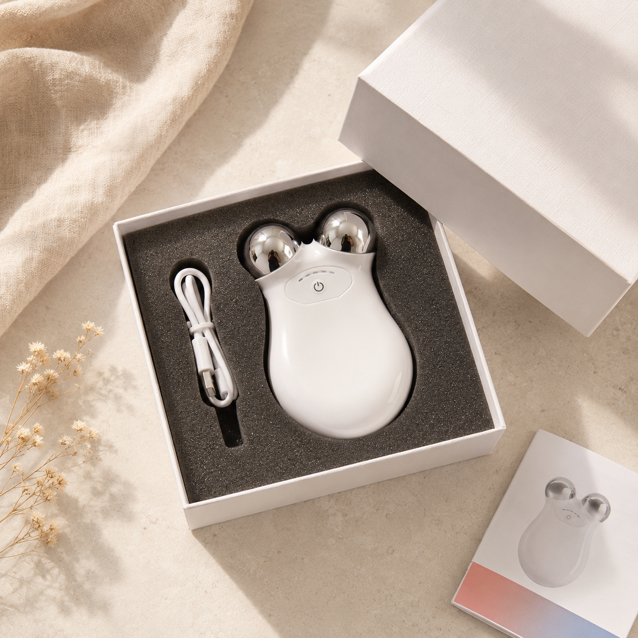 A white handheld beauty device with two silver metal ball tips, shown in its open white gift box with a charging cable and dark foam insert. The box is styled on a light beige surface with a draped fabric and small dried flowers.