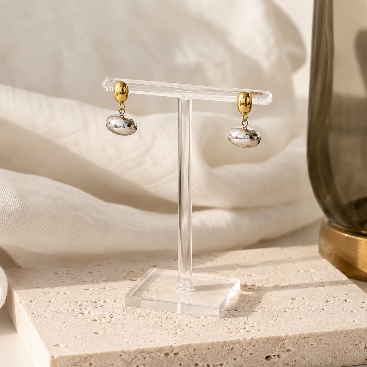 A pair of dangling earrings displayed on a clear acrylic stand. Each earring has a polished gold oval stud connected by a small chain to a silver oval drop.