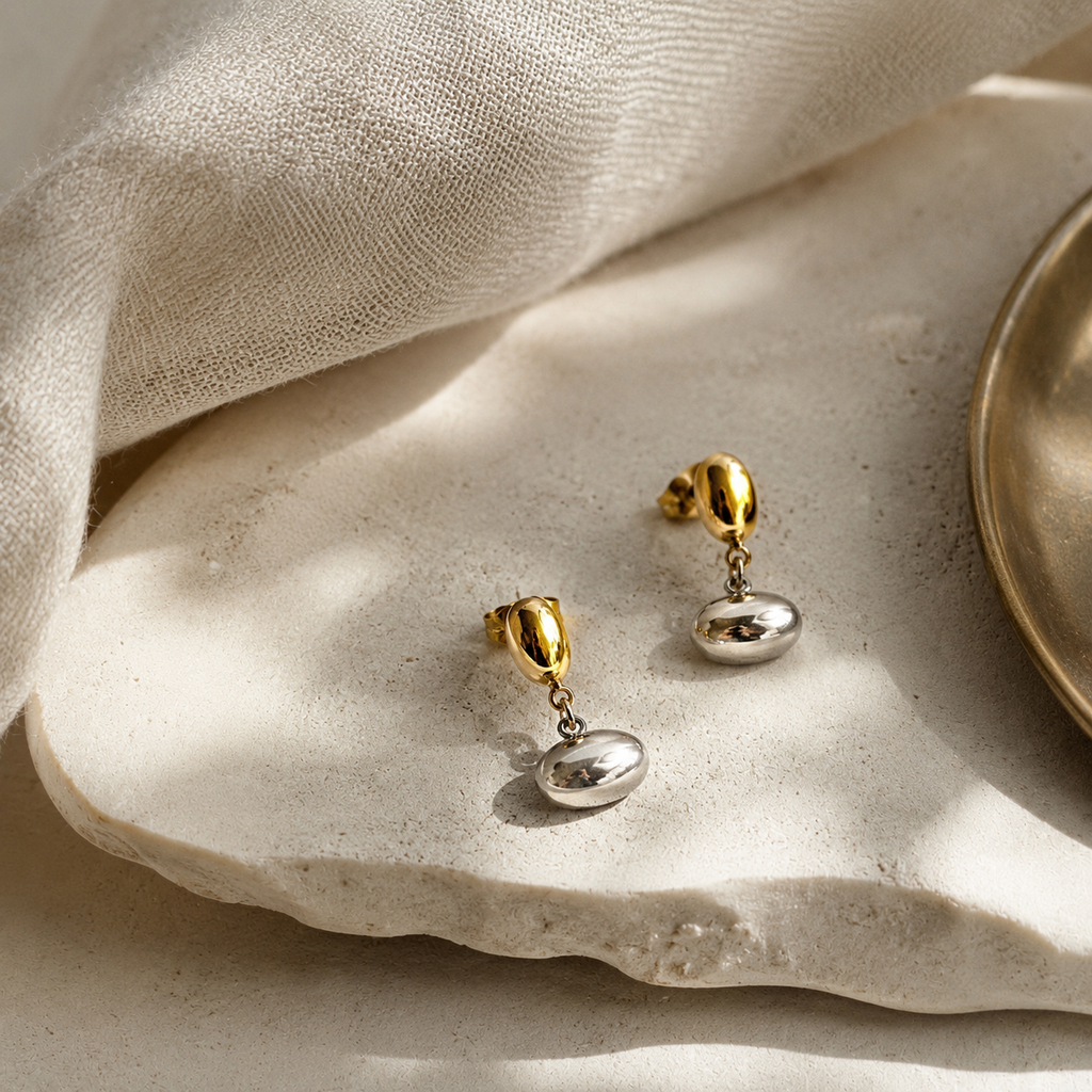 Two small drop earrings with gold posts and polished silver oval charms, arranged on a light stone surface with draped fabric and a partial metallic dish in the background.