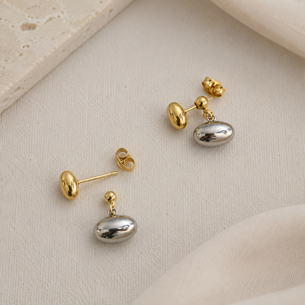A pair of gold and silver drop earrings with polished oval silver pendants and gold posts, displayed on a textured beige fabric background.