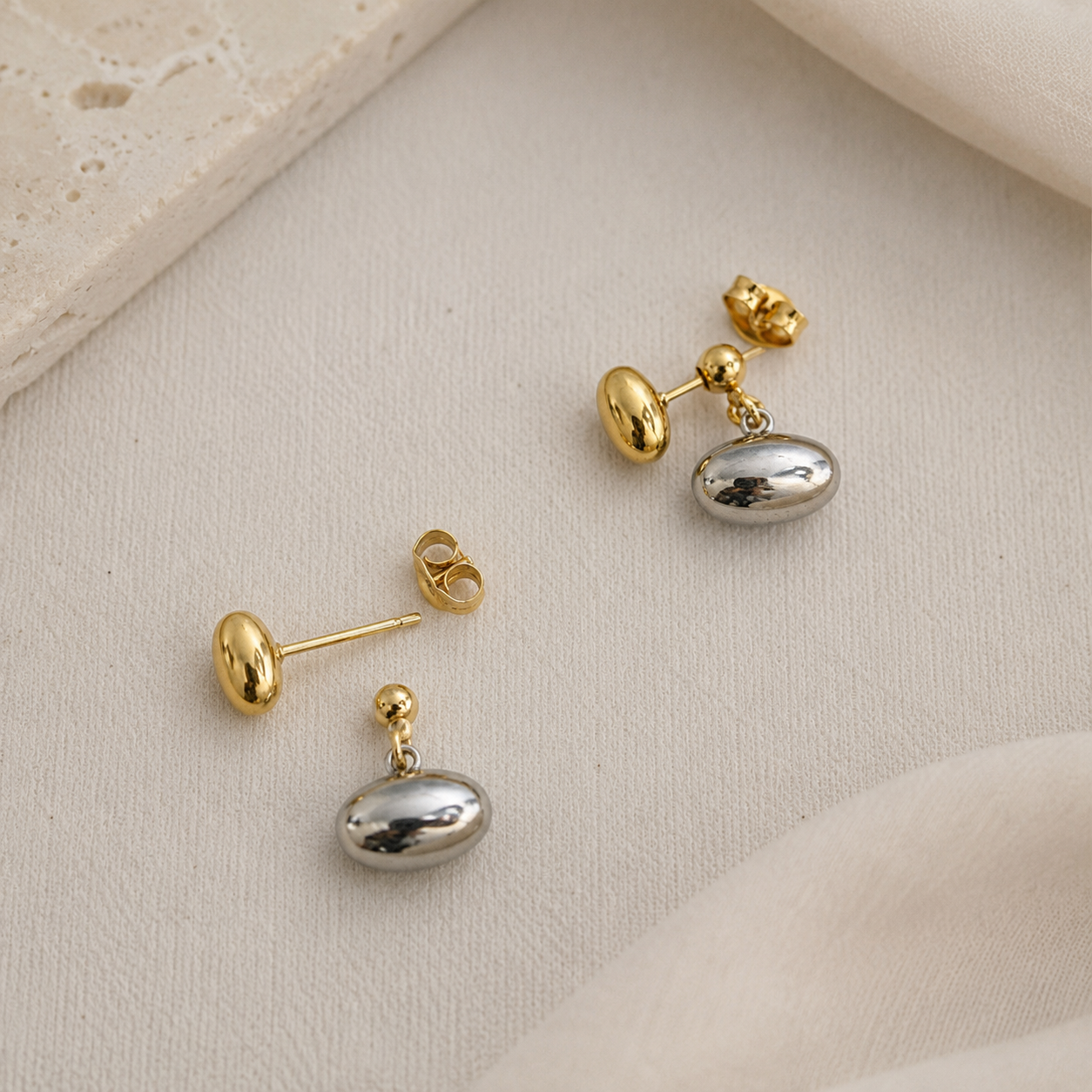 A pair of gold and silver drop earrings with polished oval silver pendants and gold posts, displayed on a textured beige fabric background.