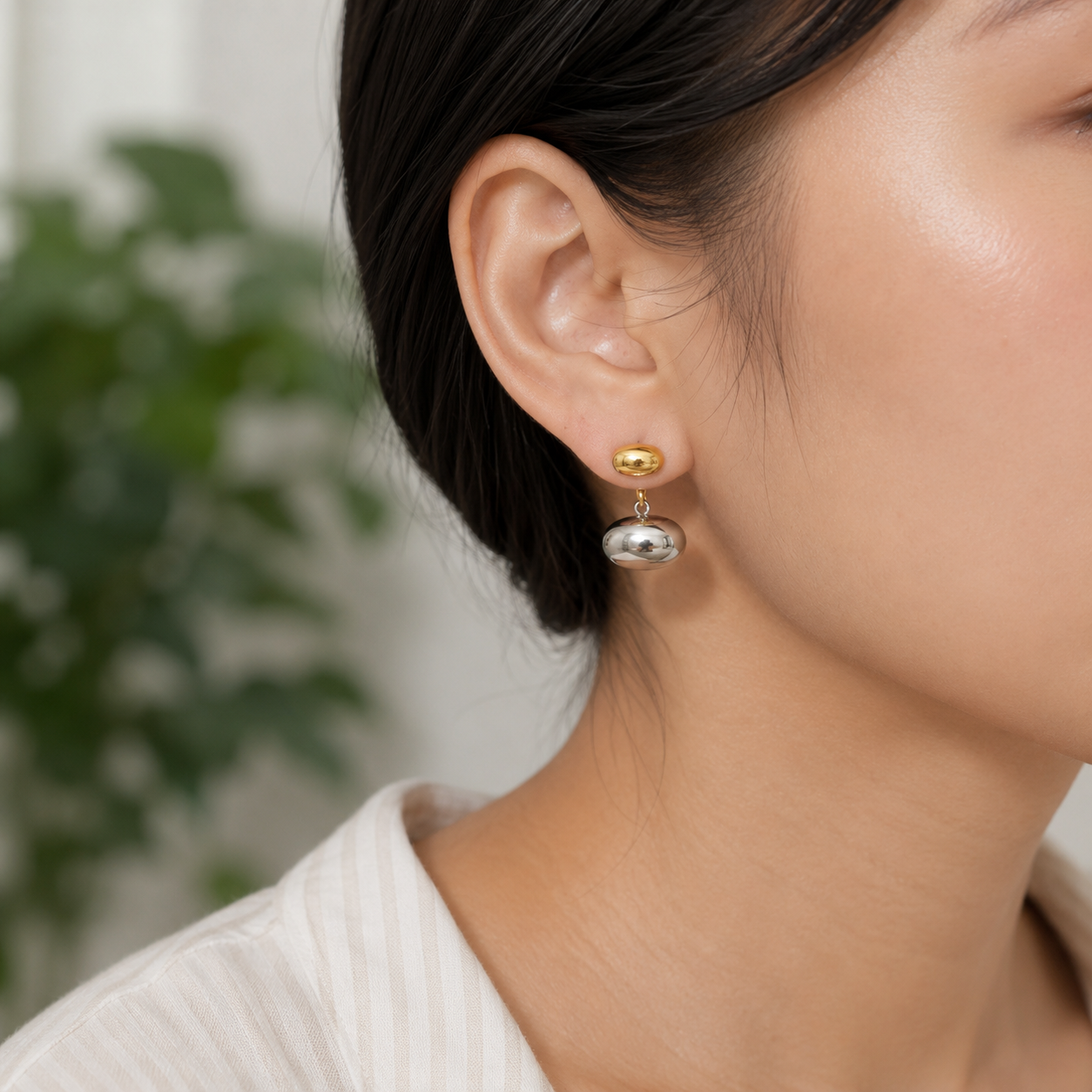 A close-up of a woman's ear wearing a small gold stud earring with a dangling polished silver disc, against a softly blurred indoor background with greenery.