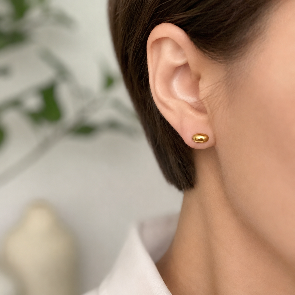 A close-up of a person's ear wearing a small, polished gold stud earring, with a soft-focus indoor background.