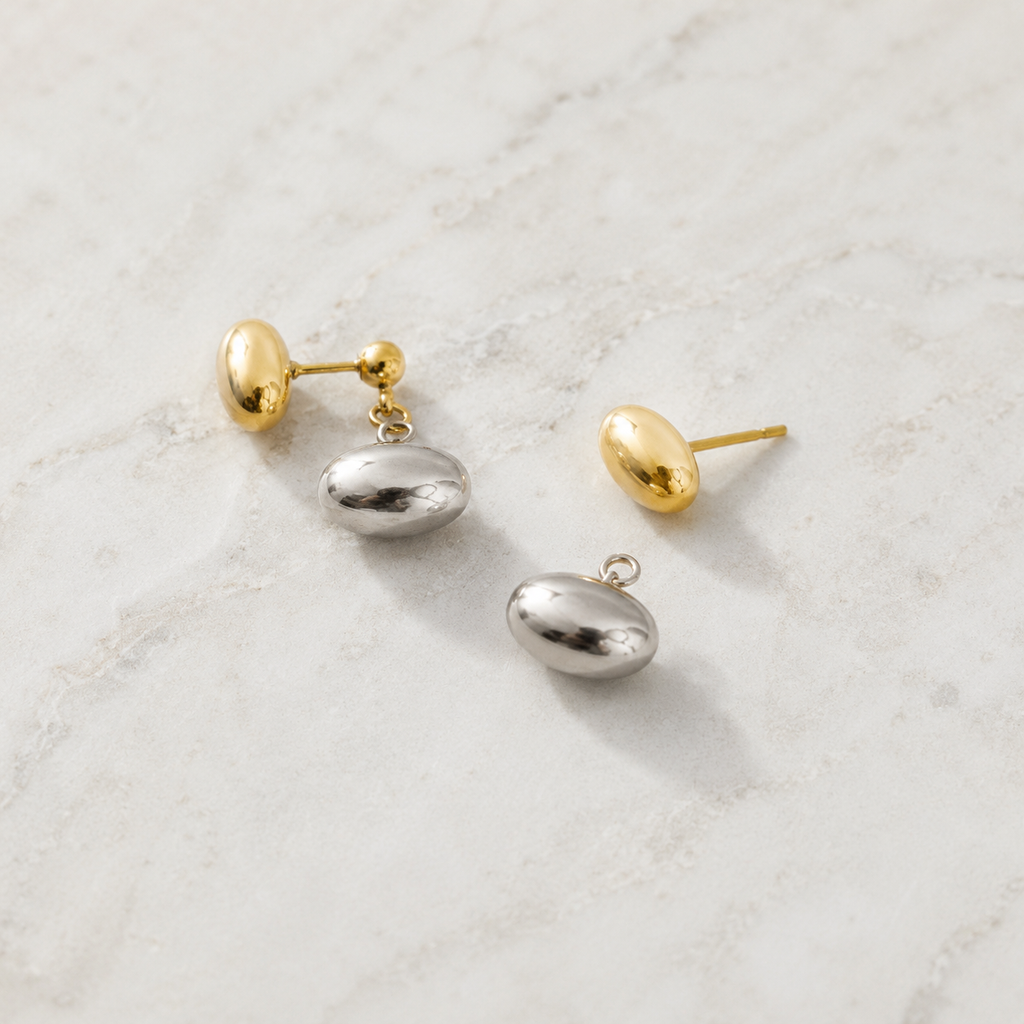 Three minimalist stud earrings with polished gold and silver oval charms, displayed on a light marble surface.