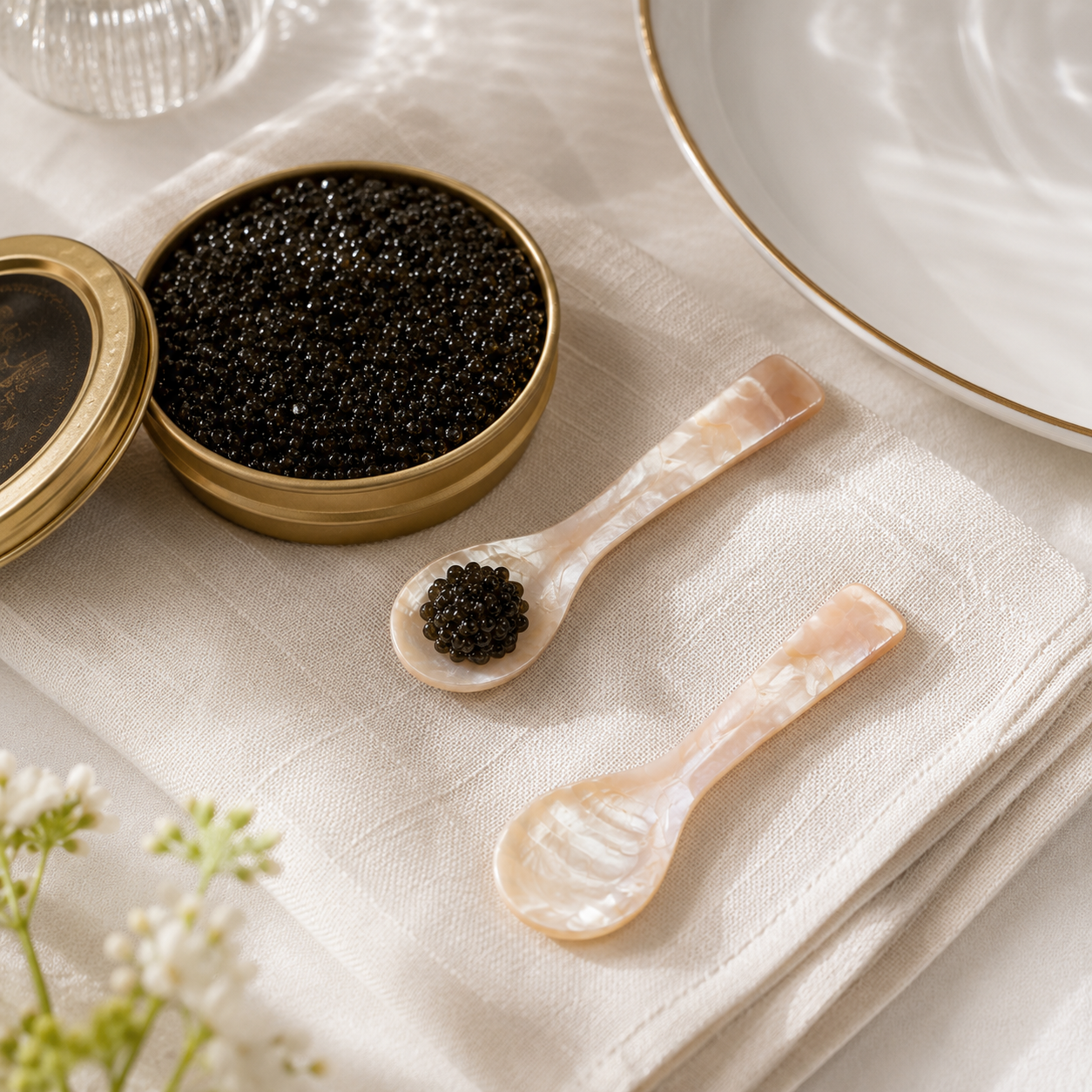 Two small pearlescent spoons resting on a cream linen napkin beside an open round gold tin filled with black caviar, with part of a white plate visible in a softly lit tabletop setting.