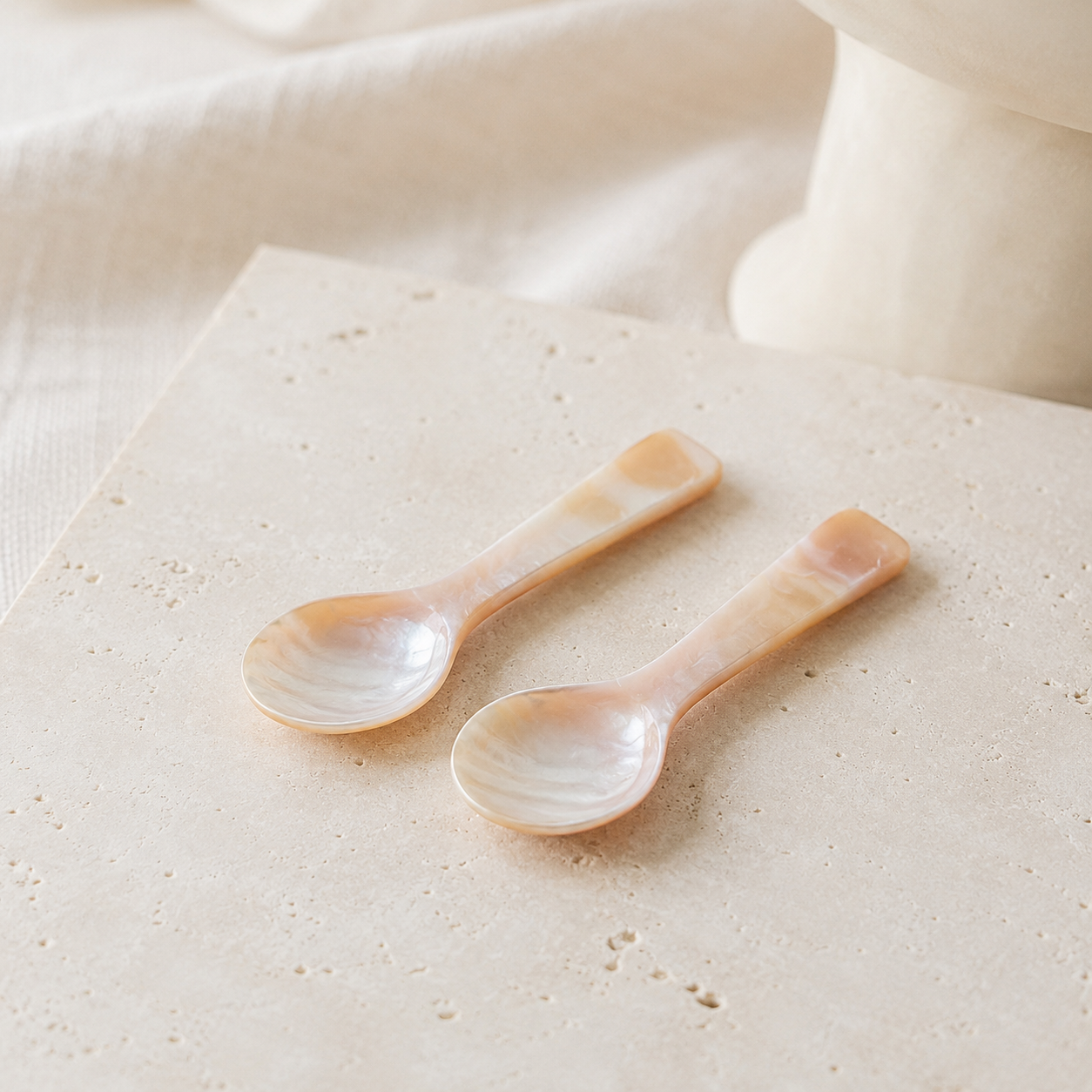 Two small shell spoons with pearlescent peach and cream tones resting on a textured beige stone surface with soft neutral fabric in the background.