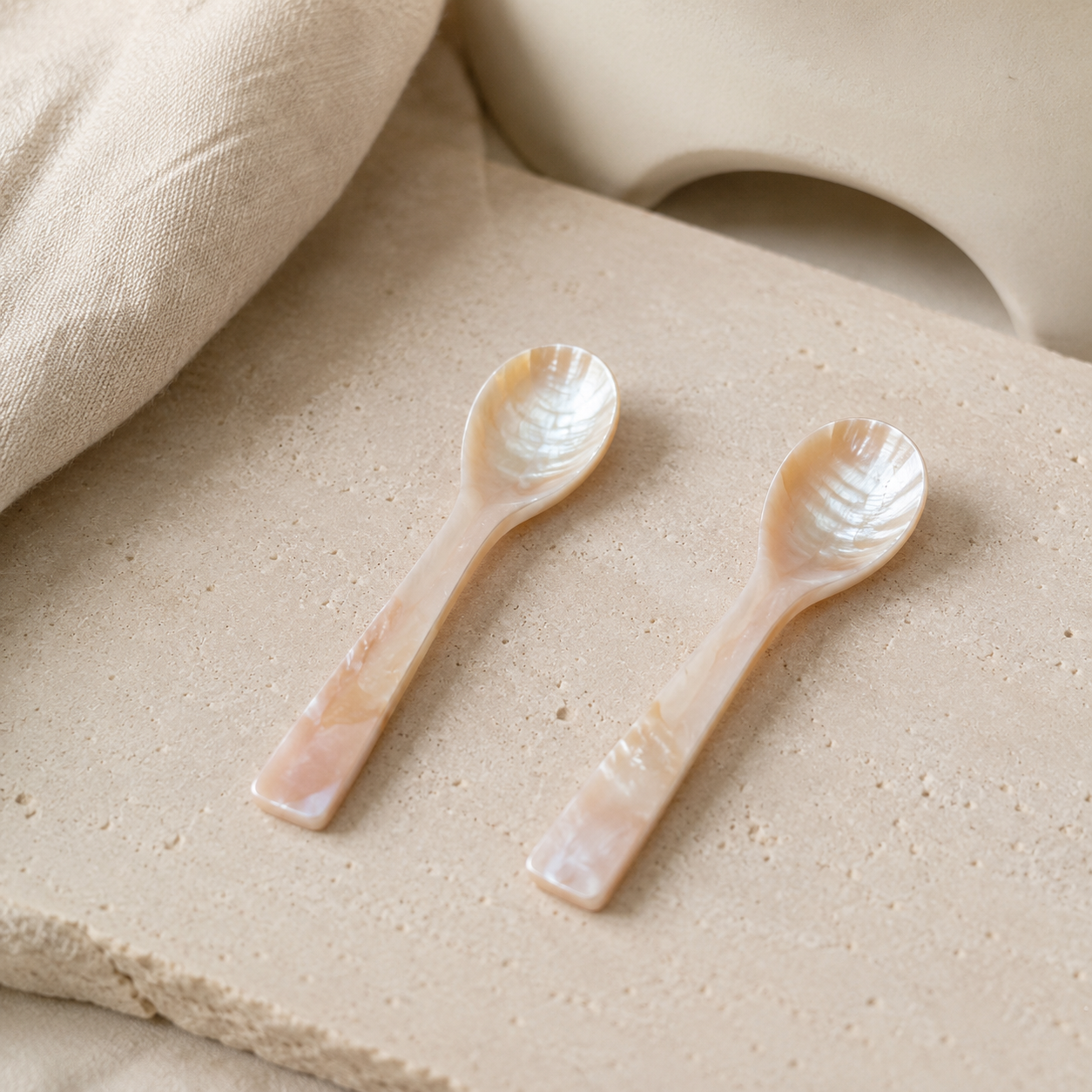 Two small mother-of-pearl spoons with a glossy, iridescent finish, resting on a textured beige stone slab with a soft beige fabric in the background.