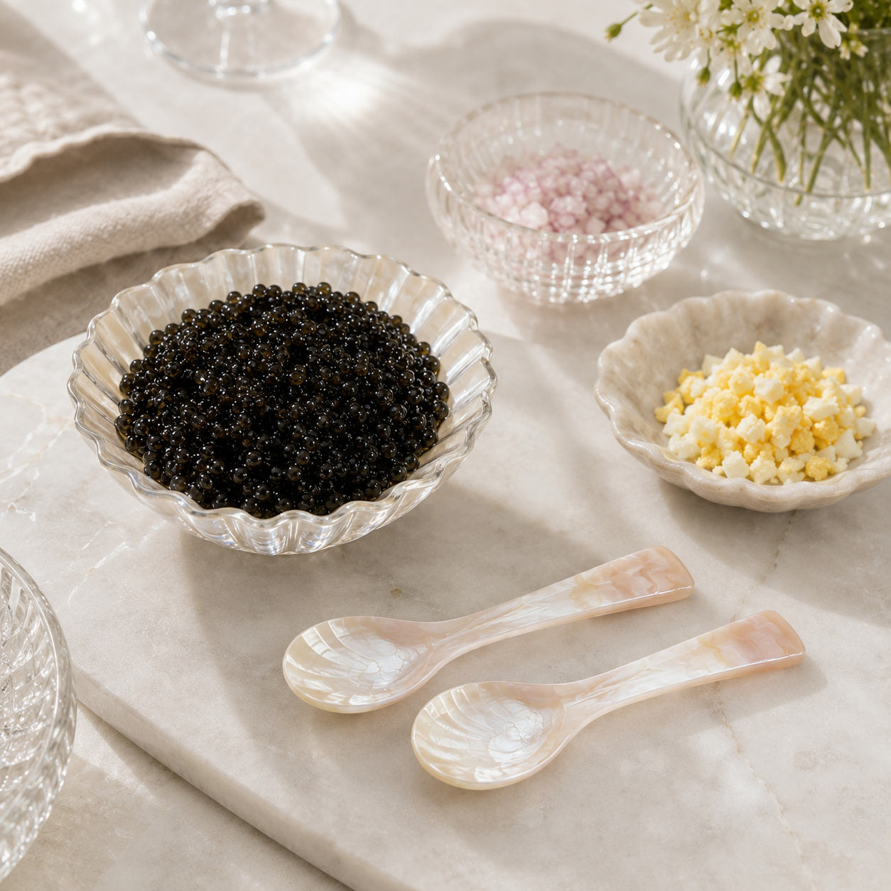 A clear scalloped glass bowl filled with black caviar on a light marble surface, alongside two small mother-of-pearl spoons, a small dish of pink salt, and a small dish of yellow cheese, with a folded beige cloth in the background.