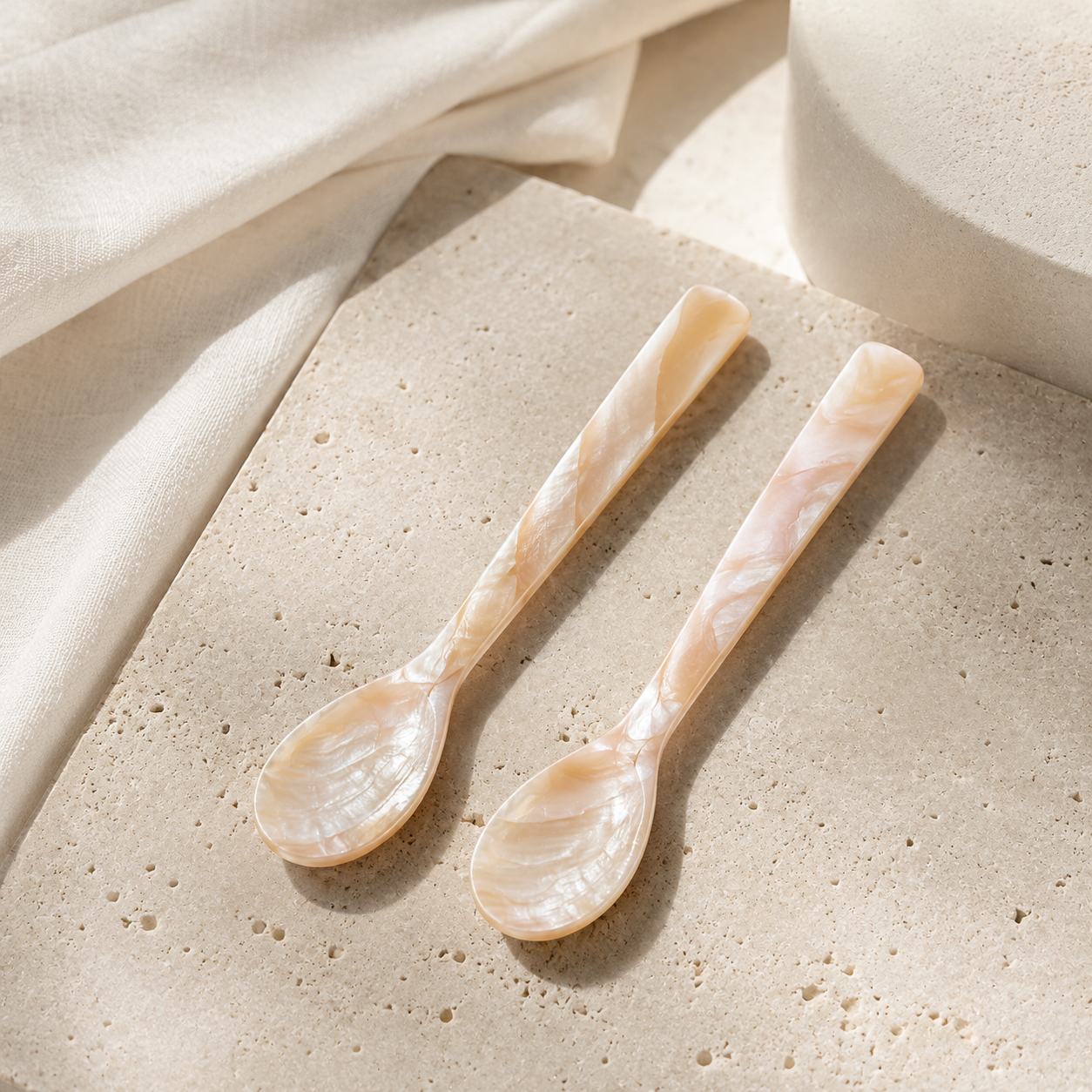Two long-handled spoons with pearlescent mother-of-pearl style handles and oval bowls, shown on a textured beige stone surface with draped cream fabric and a soft neutral prop in the background.