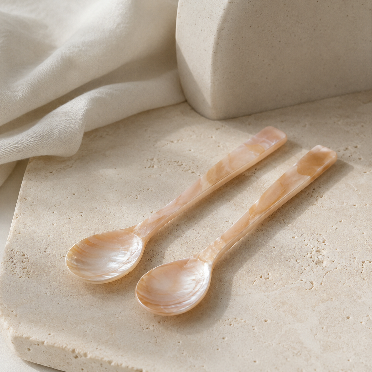 Two small mother-of-pearl spoons with a warm, iridescent beige and cream color, resting on a textured beige stone surface next to a draped white fabric.