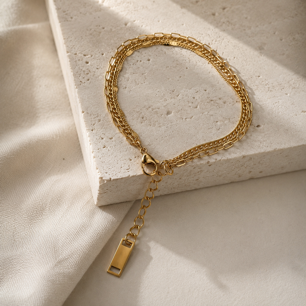 A gold chain bracelet with a lobster clasp and a dangling rectangular tag, styled on a textured white stone block with soft cream fabric in the background.