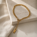A gold chain bracelet with a lobster clasp and a dangling rectangular tag, styled on a textured white stone block with soft cream fabric in the background.
