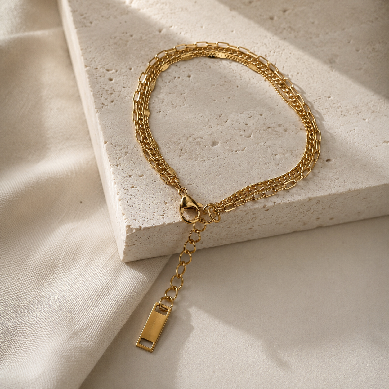 A gold chain bracelet with a lobster clasp and a dangling rectangular tag, styled on a textured white stone block with soft cream fabric in the background.
