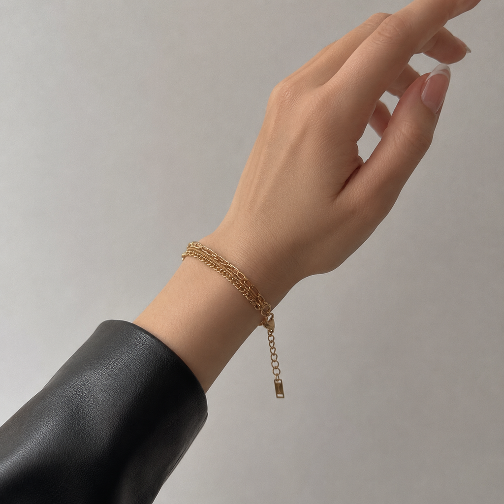 A delicate gold chain bracelet with an adjustable lobster clasp and small dangling tag, shown worn on a person's wrist against a plain light gray background.