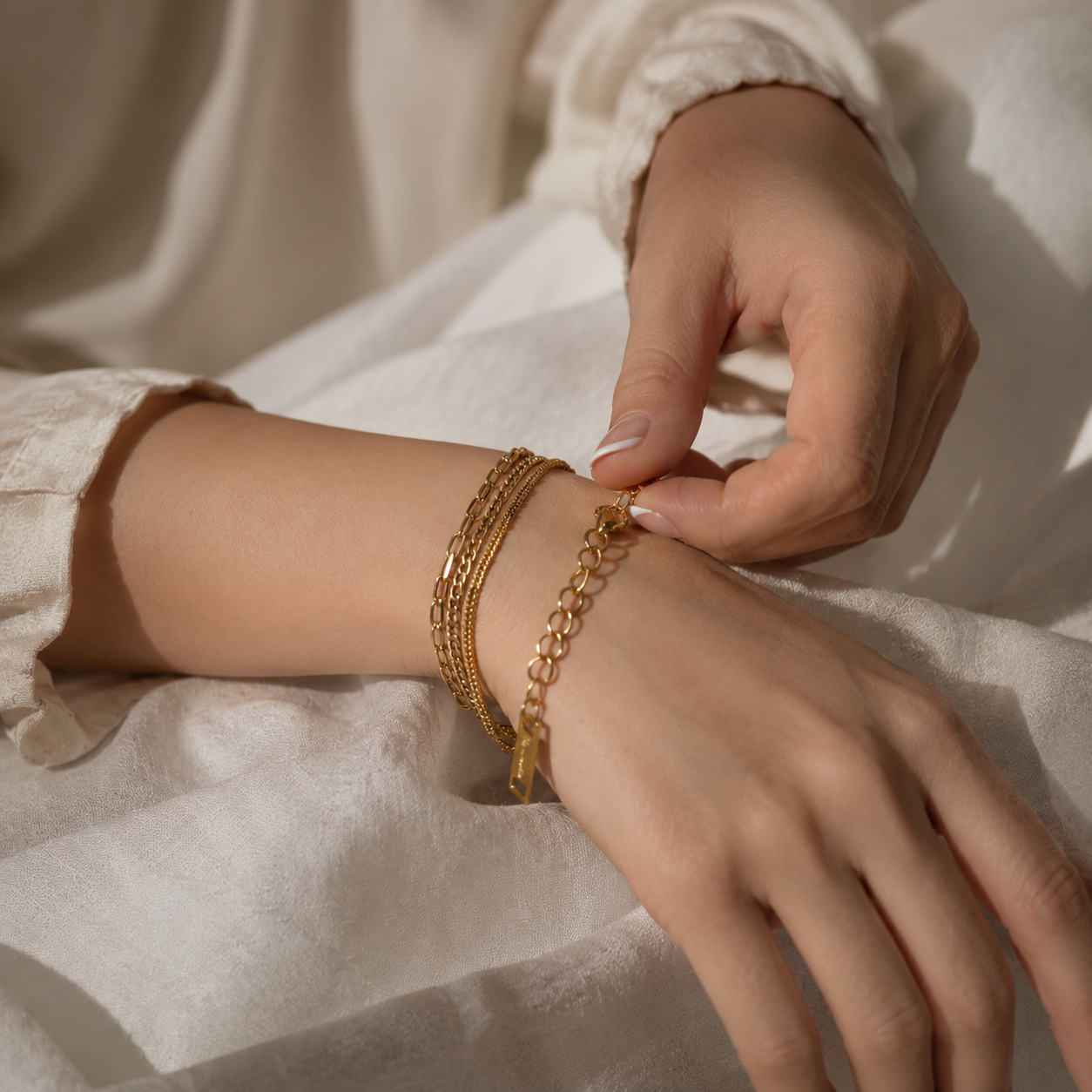 A close-up of a person's hands and wrist wearing a stack of gold chain bracelets, including a delicate nameplate bracelet and a textured chain bracelet, resting on a soft white linen fabric.