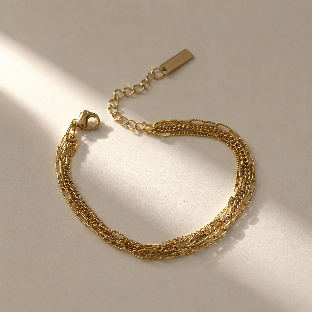 A gold-toned multi-strand chain bracelet with a lobster clasp and extension chain, photographed on a light beige surface with soft diagonal shadows.