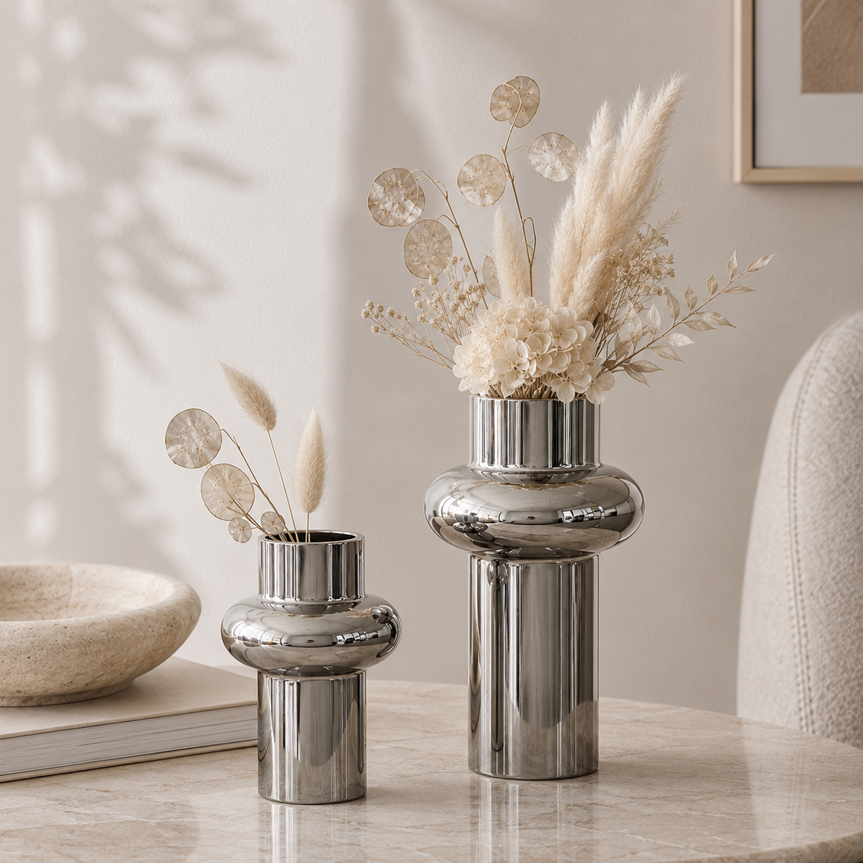 Two polished silver ceramic vases with a rounded, tiered design, one small and one large, styled with dried pampas grass and round eucalyptus leaves on a light marble tabletop in a softly lit interior setting.