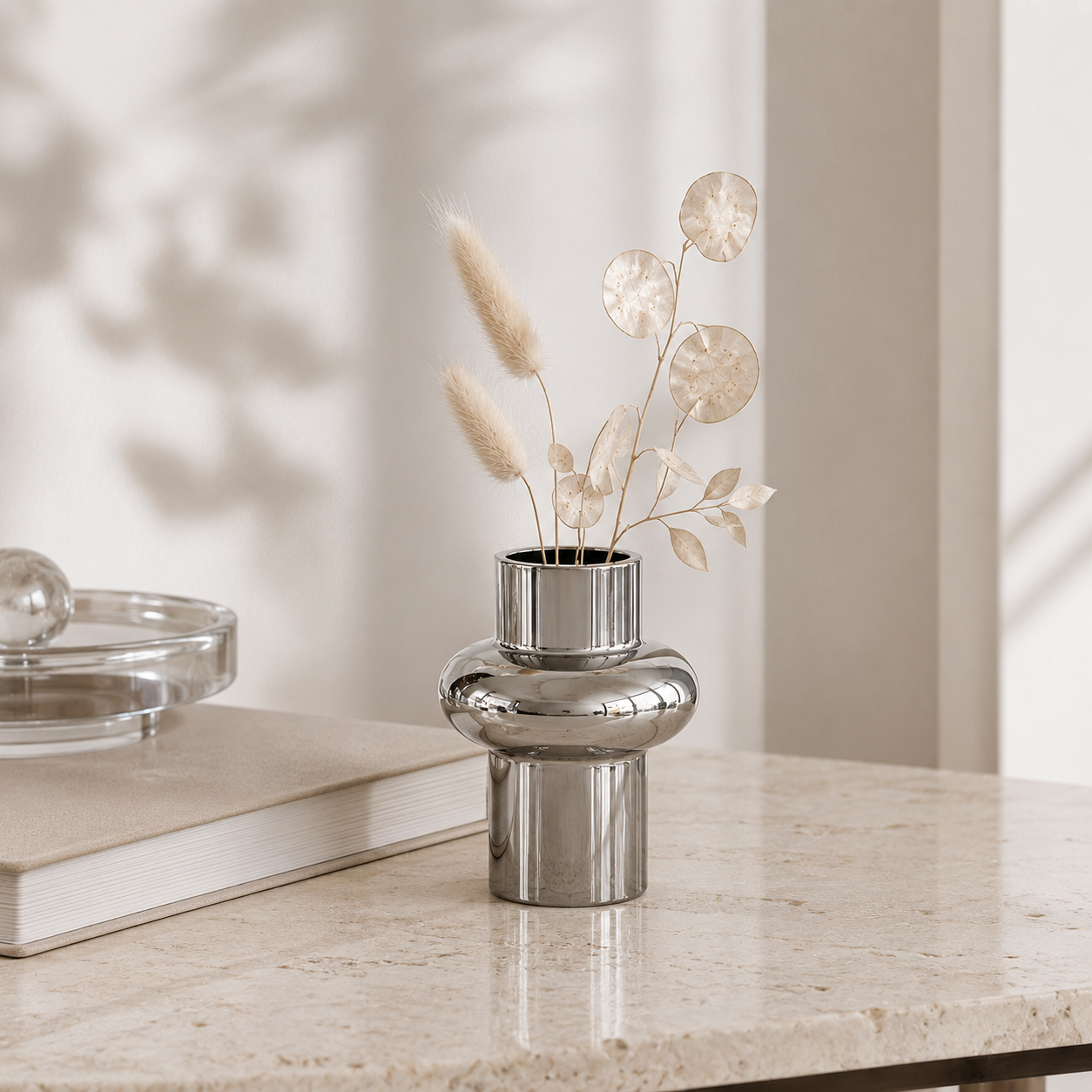 A polished silver metallic vase with a flared cylindrical top and a rounded middle section, holding dried beige grass and leaf stems, set on a light stone tabletop beside stacked books.