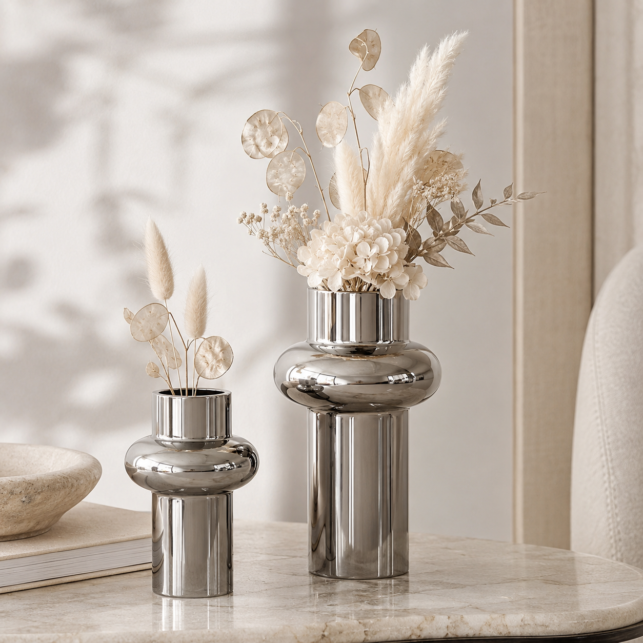 Two polished silver ceramic vases with rounded, bulbous bodies and cylindrical legs, styled with dried pampas grass and cream flowers on a light marble surface in a softly lit interior setting.