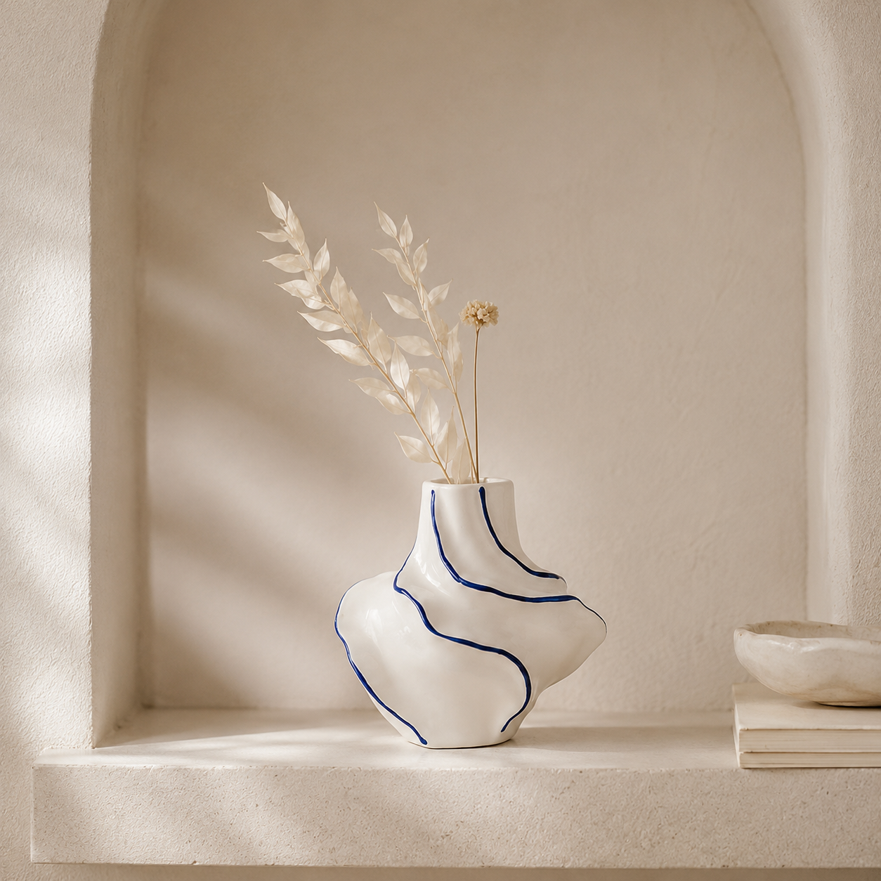 A sculptural white ceramic vase with organic blue line details and dried botanical stems, displayed on a textured stone shelf against a warm beige arched wall.