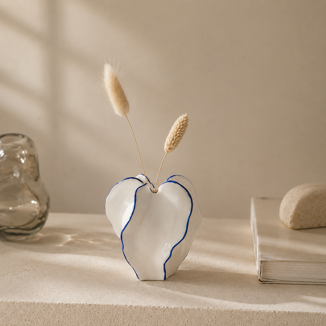 A white ceramic vase with a heart-like organic shape and thin blue line accents, holding two dried grass stems, styled on a beige surface with soft shadows and a glass object in the background.