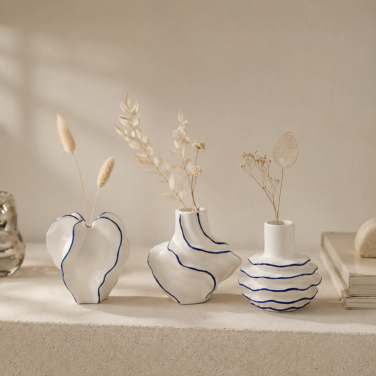 Three white ceramic vases with blue line details, arranged on a beige stone surface with dried grass stems, against a warm beige background.