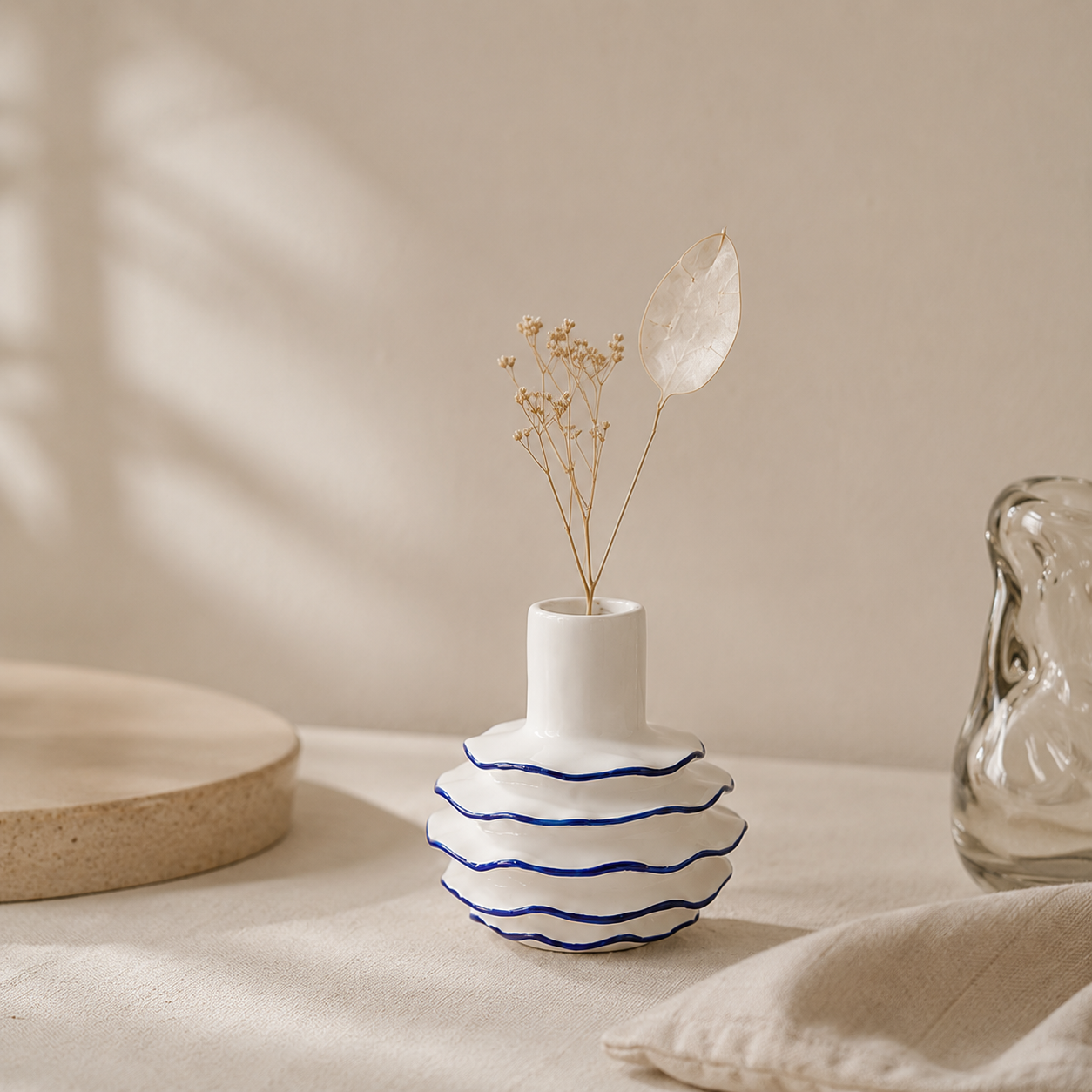 A white ceramic vase with a flared cylindrical neck and a rounded body featuring wavy blue stripes, holding dried flowers, styled on a beige fabric surface with soft shadows.