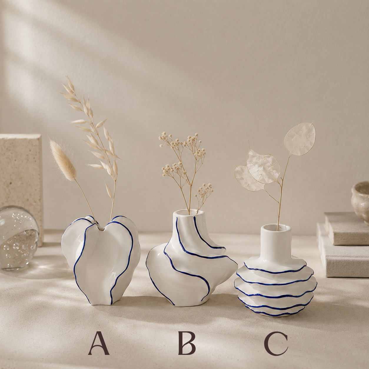 Three white ceramic vases with blue line details, labeled A, B, and C, arranged on a beige surface with dried botanical stems. The background is a warm beige wall with soft shadows.