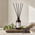 An amber glass reed diffuser bottle with black reed sticks, displayed on a rustic wooden board with eucalyptus leaves, lavender sprigs, and a beige linen cloth against a soft neutral background.