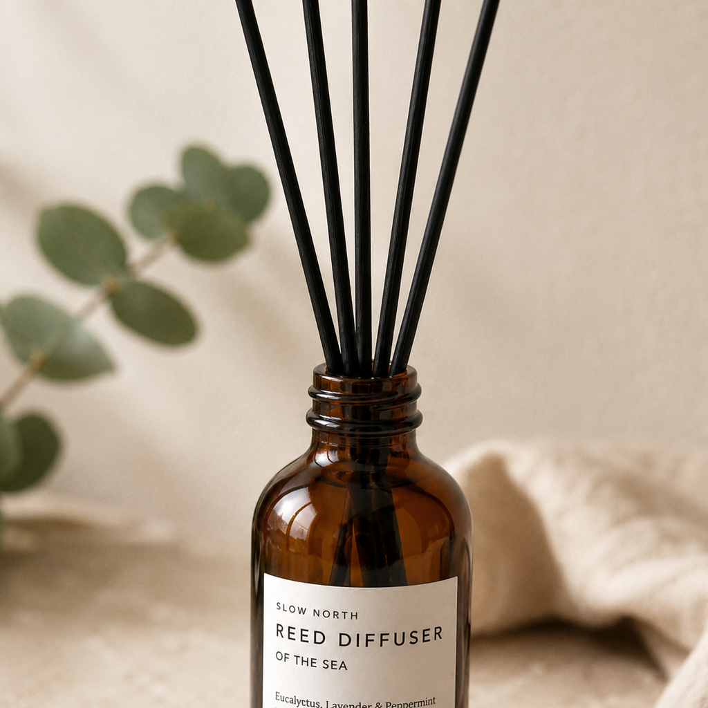 An amber glass reed diffuser bottle with black reed sticks and a white label reading 'SLOW NORTH REED DIFFUSER OF THE SEA', styled with a blurred eucalyptus branch and beige fabric in the background.