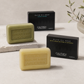Three Calithea olive oil soap bars in different shades of green and yellow-beige are displayed alongside their black packaging boxes on a light stone surface.