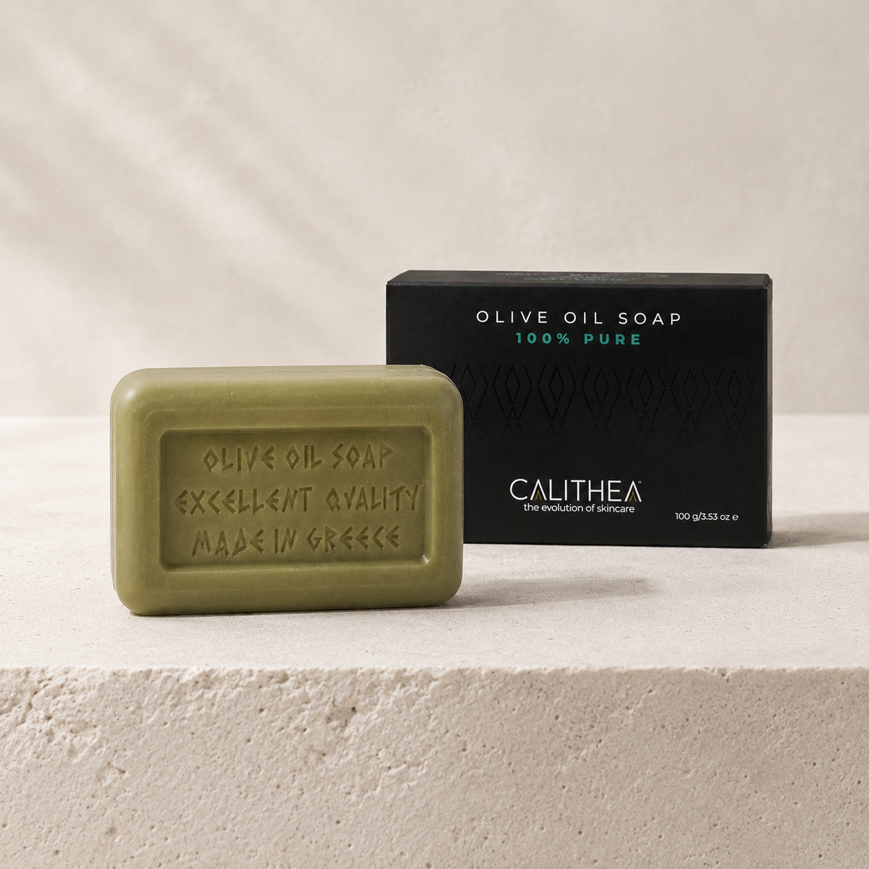 An olive green rectangular bar of Calithea olive oil soap sits in front of its black packaging box on a light beige textured stone surface.
