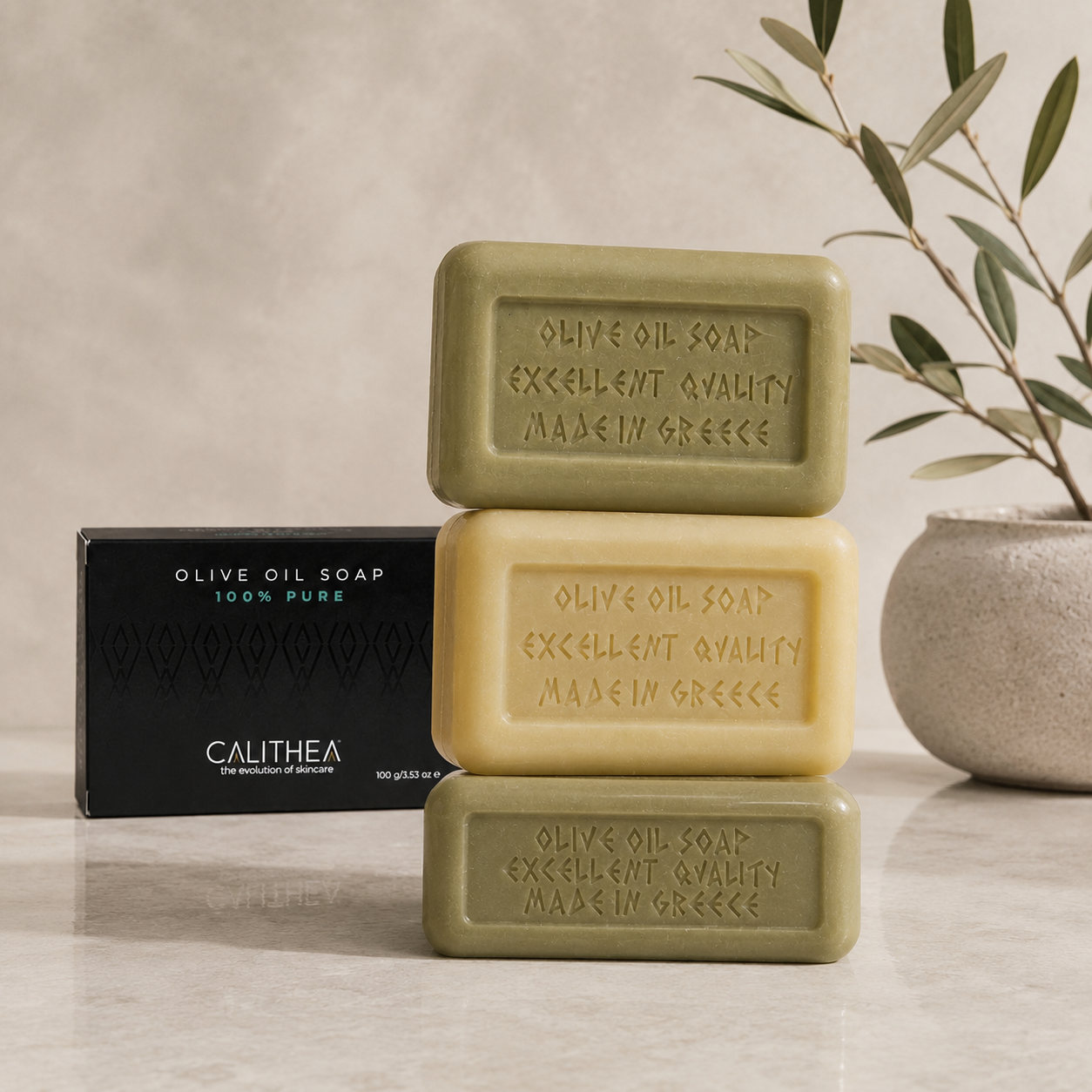 Three rectangular bars of olive oil soap stacked vertically, with a black Calithea brand box on the left and a potted olive branch on the right, set on a light marble surface against a textured beige wall.