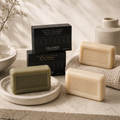 Three Calithea olive oil soap bars in different shades of green and beige, arranged on a stone surface with their black branded boxes behind them. The scene includes a ceramic bowl, dried stems, and a textured cloth in a softly lit bathroom-like setting.