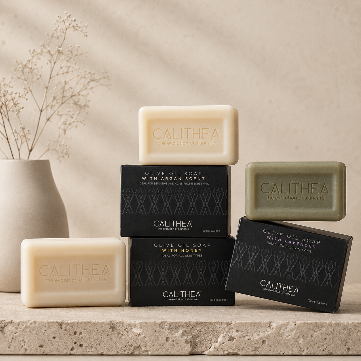 Five Calithea olive oil soap bars in different scents, displayed with their black packaging boxes on a stone ledge next to a ceramic vase with dried flowers.