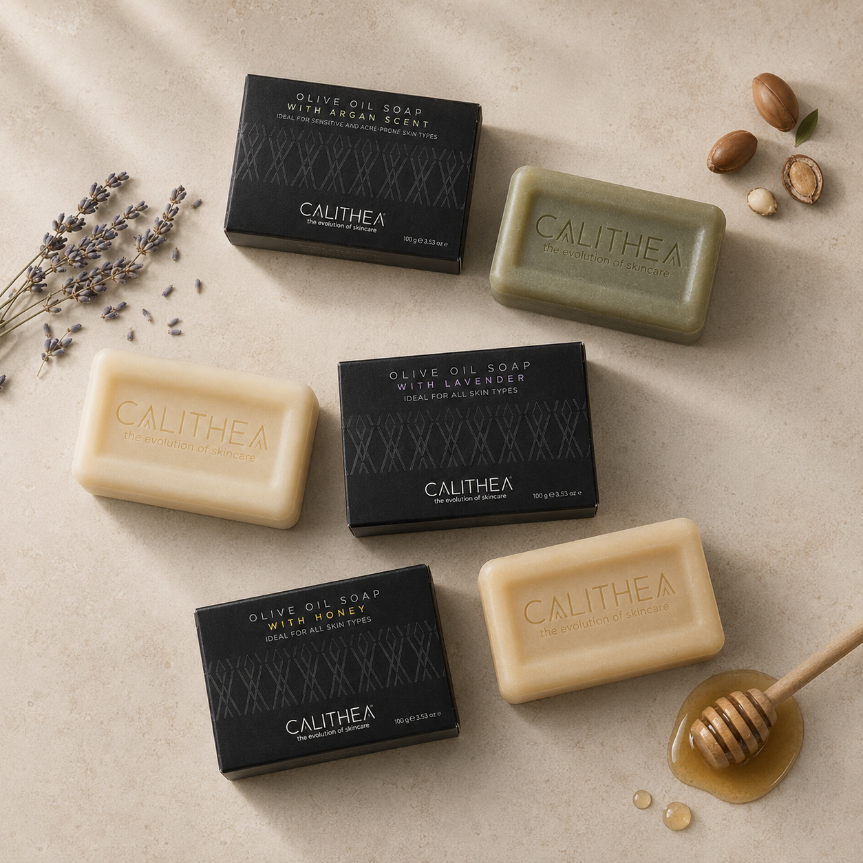 Four Calithea olive oil soap bars arranged on a light beige surface with their black boxes, plus scattered lavender sprigs, nuts, honey, and a wooden honey dipper. The soaps are shown in two tones: one pale cream and one muted olive green.