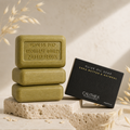 Three olive green bars of Calithea Olive Oil Soap stacked on a stone slab next to a black product box, styled with dried oat grass on a warm beige background.