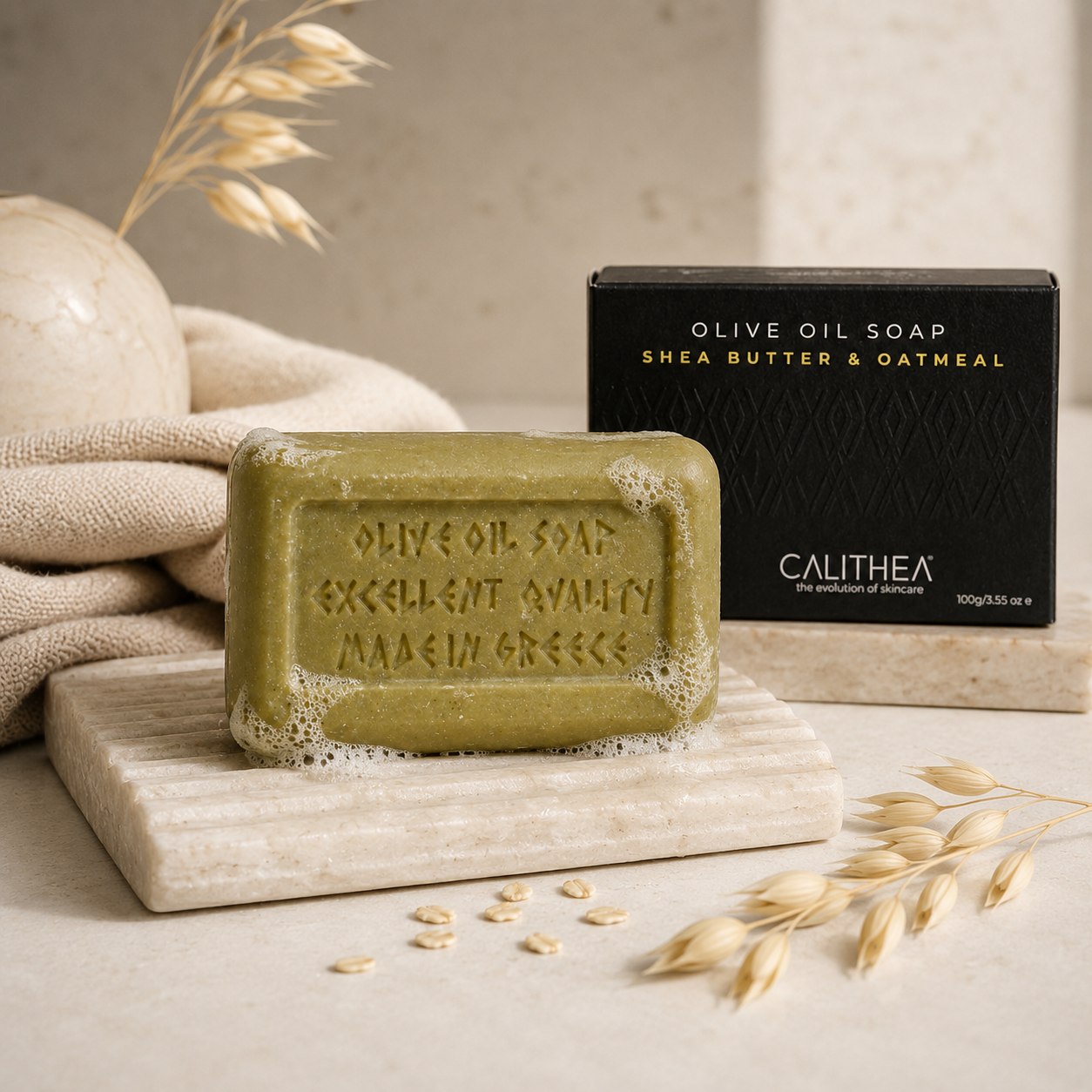 A rectangular bar of olive oil soap with a foamy edge sits on a light stone slab, with the black Calithea product box behind it and oat stems and neutral props around it.