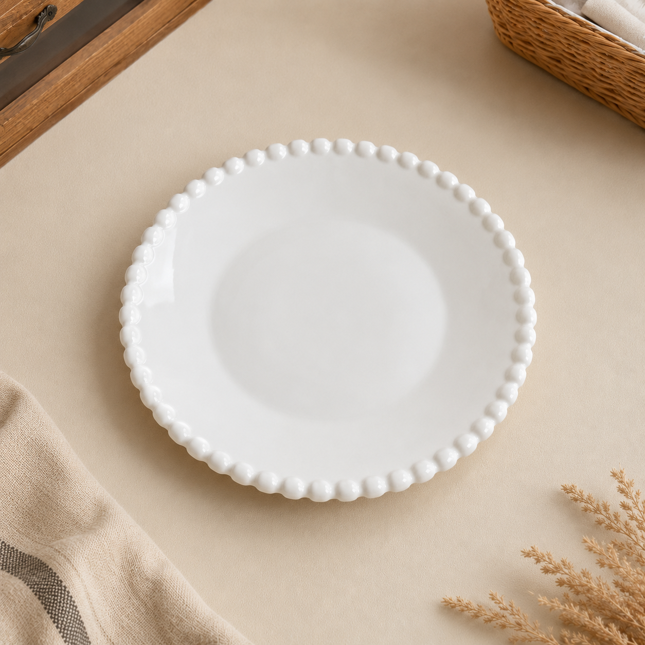 A round white ceramic plate with a decorative beaded rim, styled on a beige tabletop with neutral-toned fabric and dried floral accents.