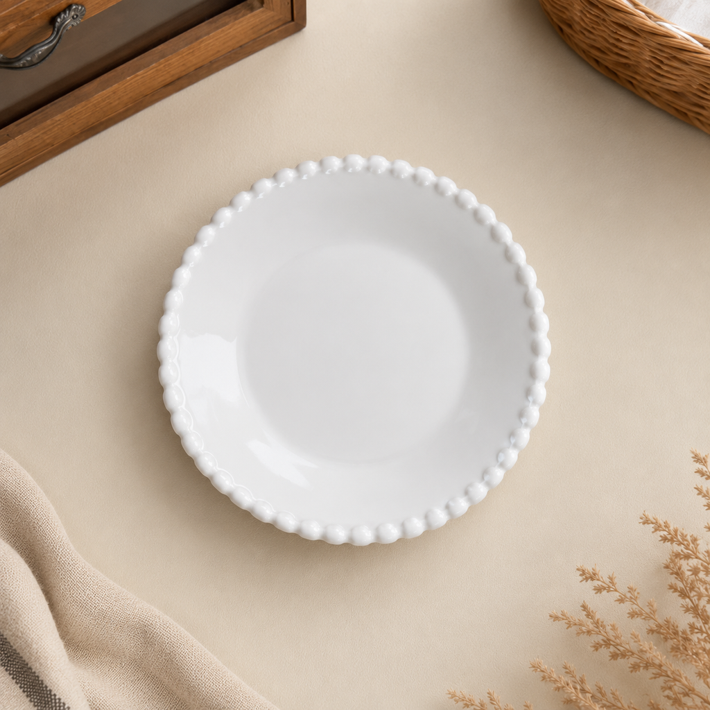 A white ceramic plate with a decorative beaded scalloped rim, photographed from above on a beige surface with soft neutral props.