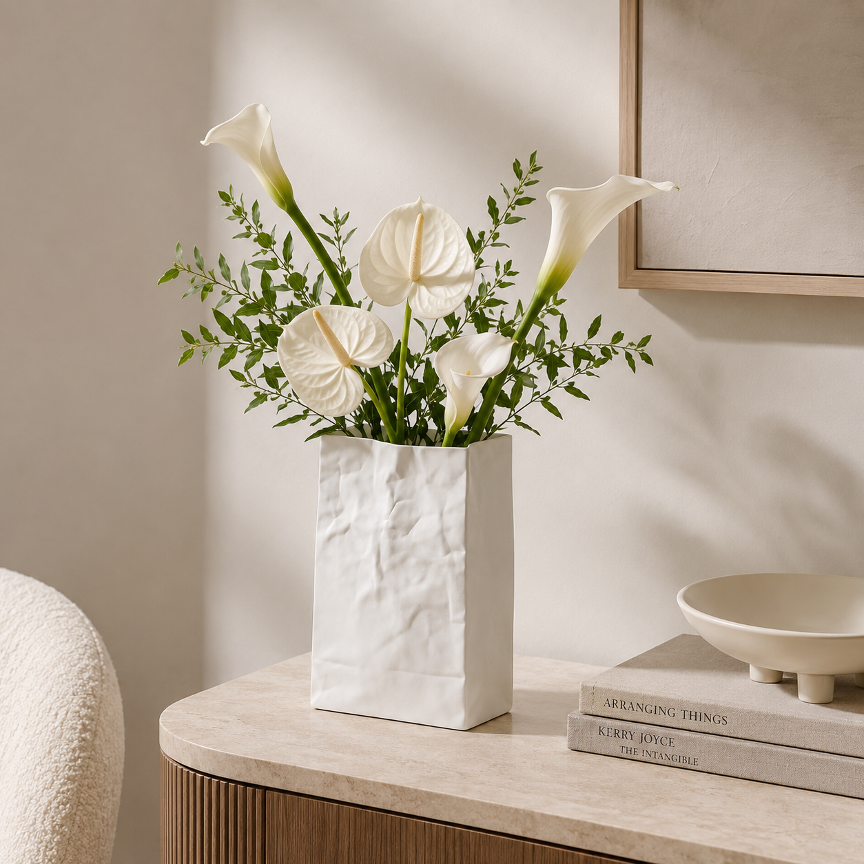 White paper bag vase filled with white calla lilies and green foliage, styled on a light stone side table in a warm neutral interior setting.