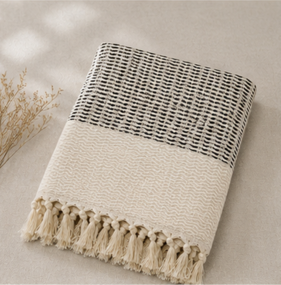 A folded woven throw blanket with a black and cream geometric pattern on the top half and a cream wavy texture on the bottom half, finished with matching tassels, displayed on a neutral beige background with dried floral stems.