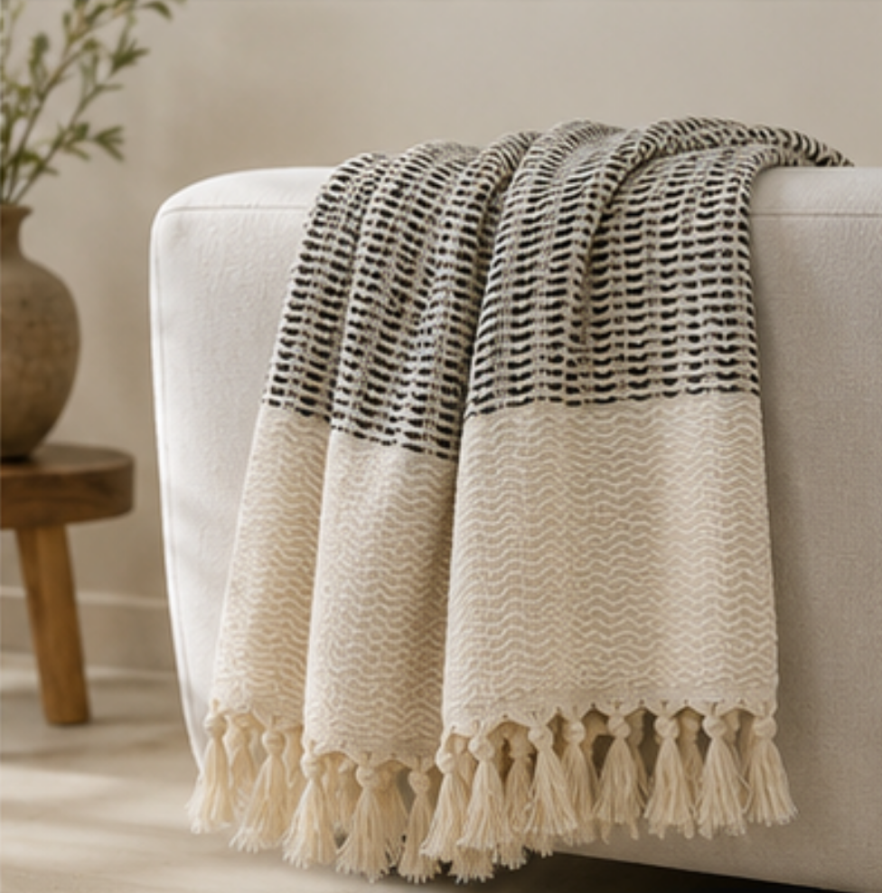 A cream and black woven throw blanket with tassels draped over a white armchair in a softly styled room.