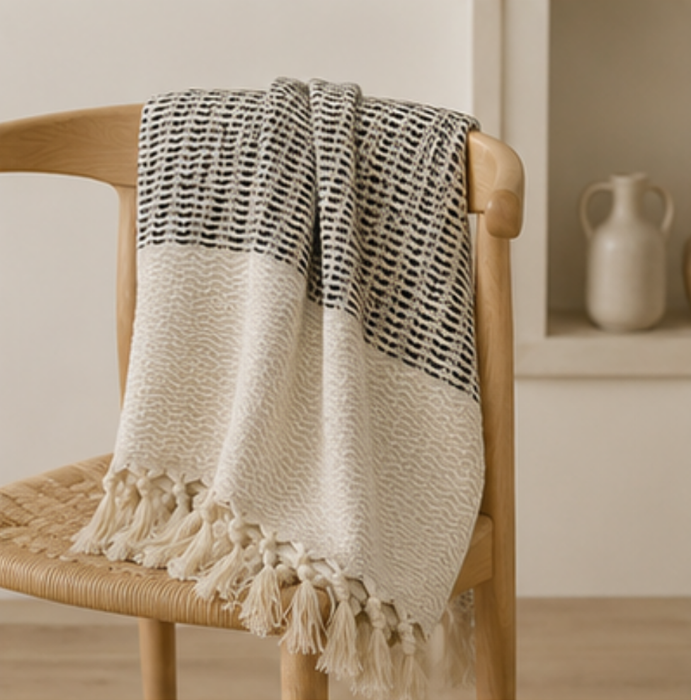A cream and black woven throw blanket with tassels draped over a light wood chair in a softly lit interior setting.