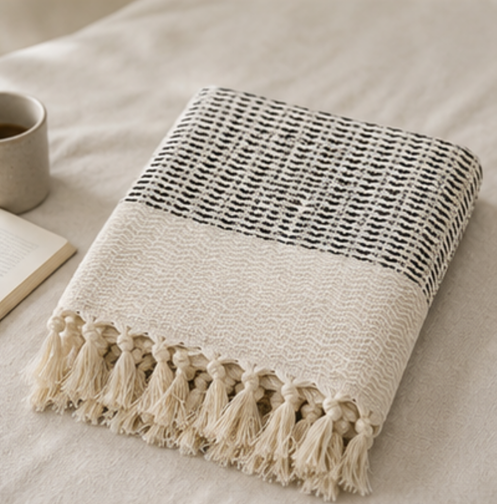 A folded woven throw blanket with cream and black stripes and tassels, styled on a light beige fabric surface next to a mug and an open book.