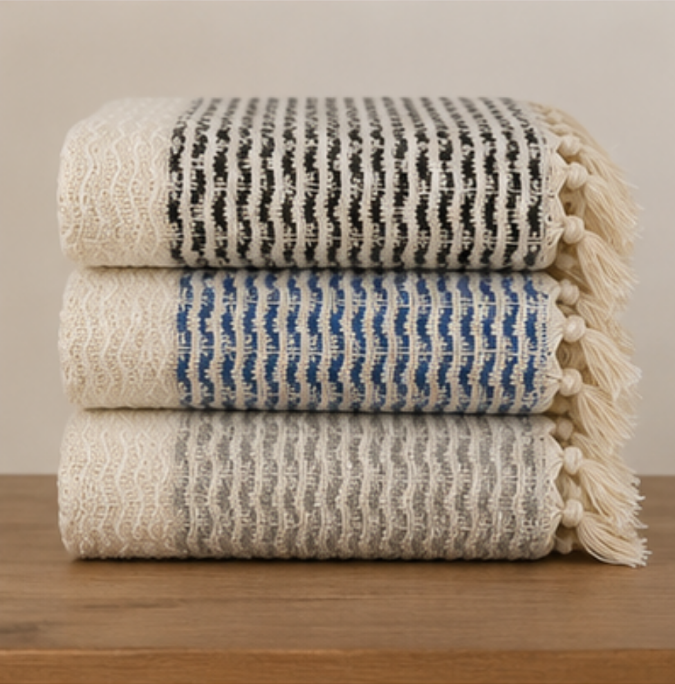 Three folded woven throw blankets stacked on a wooden surface against a plain beige wall. The top blanket is cream and black, the middle is cream and blue, and the bottom is cream.