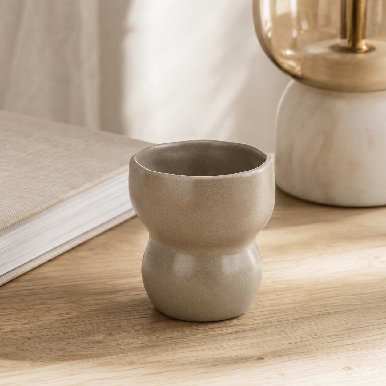 A small matte ceramic cup or candle holder with a rounded, bulbous shape sits on a light wood tabletop next to a book, with a softly blurred lamp in the background.