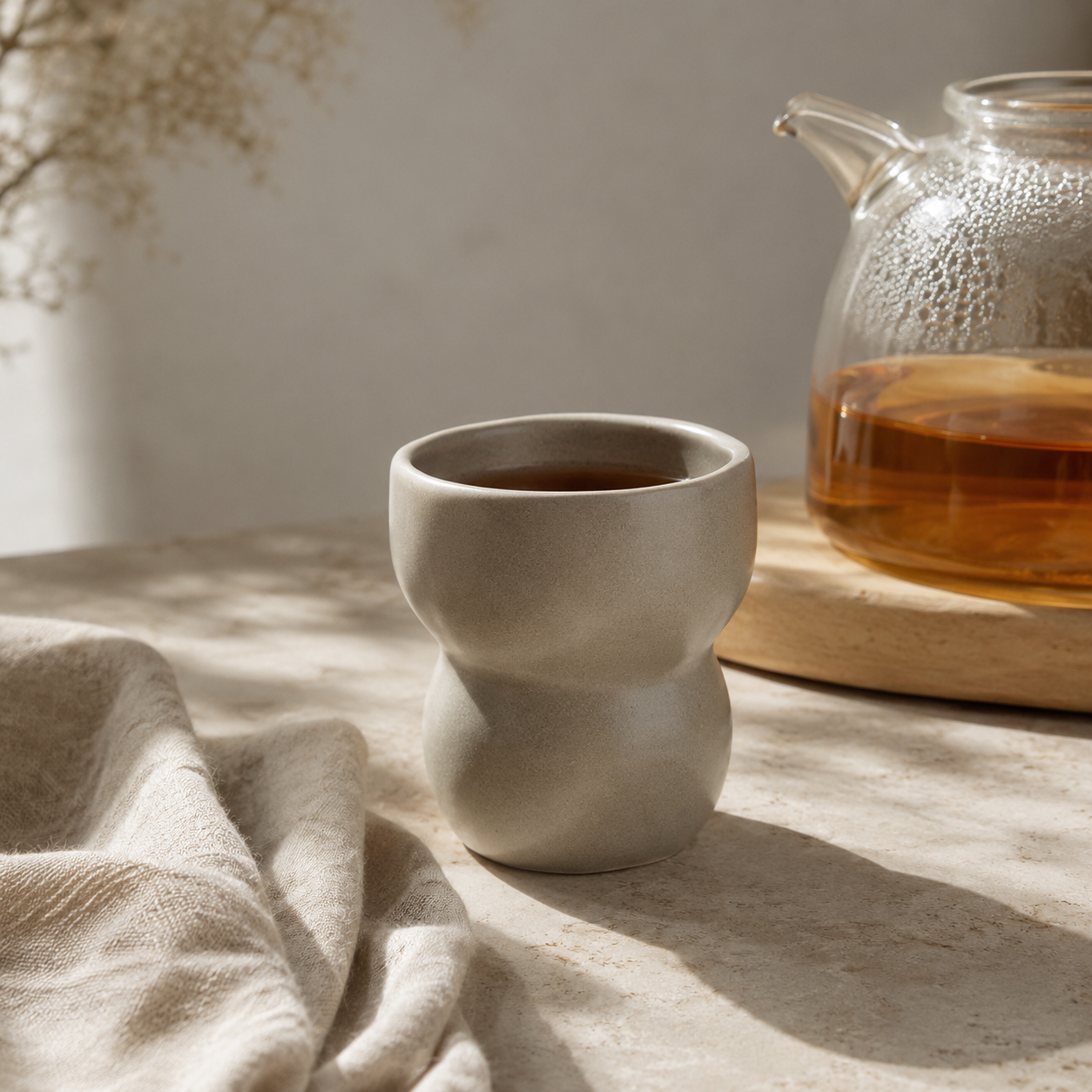 A small matte ceramic cup with a rounded, bulbous design sits on a light stone surface beside a glass teapot and draped beige fabric in a softly lit interior setting.