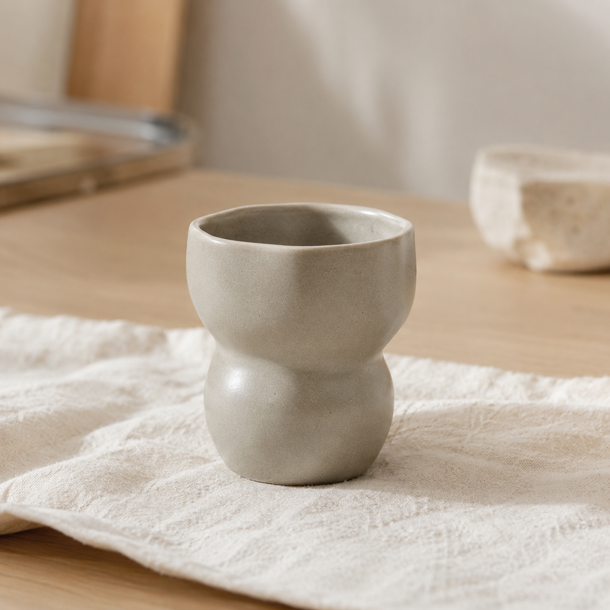 A small matte ceramic cup with a rounded, two-bulb shape sits on a white textured cloth on a light wood table.