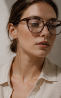 A close-up of a woman wearing tortoiseshell eyeglasses with a cream-colored collared shirt, set against a warm, neutral background.